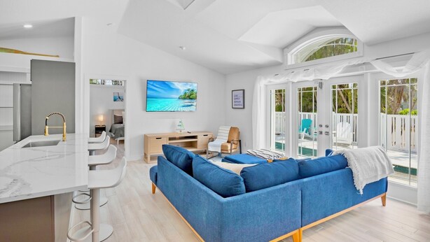 Uma sala de estar moderna com um sofá azul, uma TV e uma área de cozinha com bancada branca e banquetas.