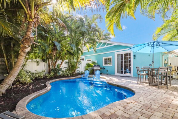 Une piscine avec une terrasse en briques, entourée de palmiers et une maison à la façade bleue.