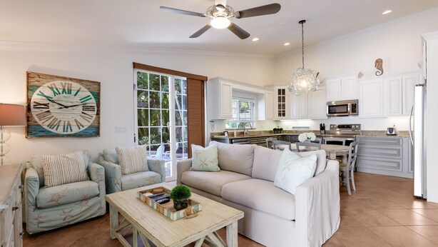 A spacious living room with a large sofa, a coffee table, and a dining area. There is a kitchen in the background with white cabinets and a ceiling fan. - Beautiful Villa near the Beach with Pool! - Mermaid's Hideaway (Holmes Beach)