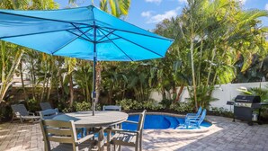 A patio with a round table, chairs, a blue umbrella, and a swimming pool surrounded by palm trees. - Beautiful Villa near the Beach with Pool! - Mermaid's Hideaway (Holmes Beach)