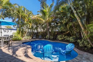 Une piscine avec deux chaises bleues, entourée de palmiers et d’un espace patio comprenant une table et des chaises.