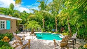 A pool area with wooden chairs, a pool, and surrounding palm trees. - Beautiful Home with Pool and Outside Living Area! Near the Beach (Anna Maria)