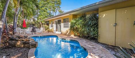 A swimming pool surrounded by a brick patio, palm trees, and a yellow building with a covered porch.