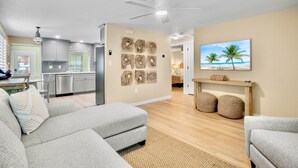 Un salon moderne avec un canapé gris, un coin repas, une cuisine et un tableau sur le thème de la plage accroché au mur.