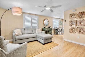 A living room with a sectional sofa, a floor lamp, a ceiling fan, and a dining area with a table and chairs. - Remodeled Duplex with Heated Pool, 2 Blocks to the Beach! (Anna Maria)