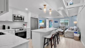 A modern kitchen with white cabinetry, a central island, and a dining area with a grey sofa and bar stools. - Duplex with Heated Pool! 2 Blocks to Beach! - Turtle Crawl (Anna Maria)
