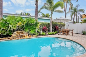 A swimming pool with a stone waterfall, surrounded by tropical plants and a patio with outdoor furniture.
