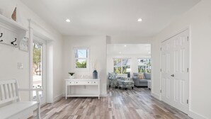 Een ruime hal met een houten vloer, een witte bank, een console tafel met een plant en uitzicht op een strand door het raam.