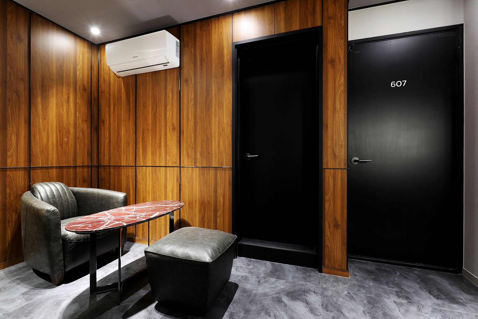A modern room with a leather armchair, a small table, a wooden panel wall, and a black door marked with the number 607.