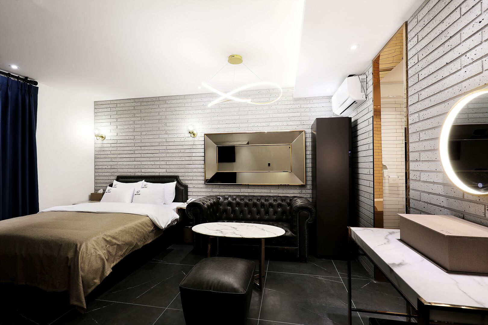 A modern hotel room with a bed, a black leather sofa, a small round table, a mirror, and a built-in air conditioning unit.