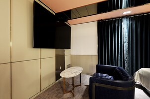 Habitación de hotel con un televisor de pantalla plana en la pared, una mesa redonda de mármol, una butaca tapizada en tono azul oscuro y una cama con cubierta blanca.
