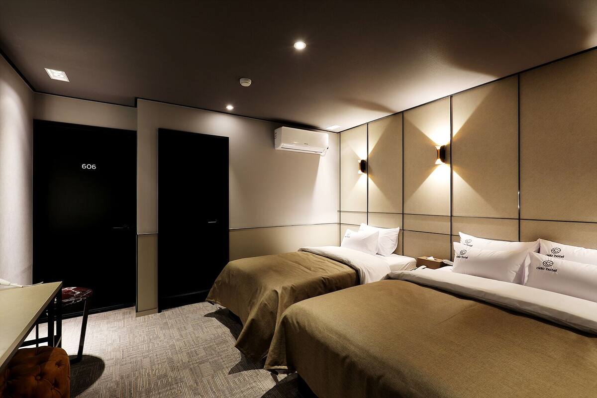 A hotel room with two beds, a desk, and a chair. There is a door with a number 606 on it, and an air conditioning unit on the wall.