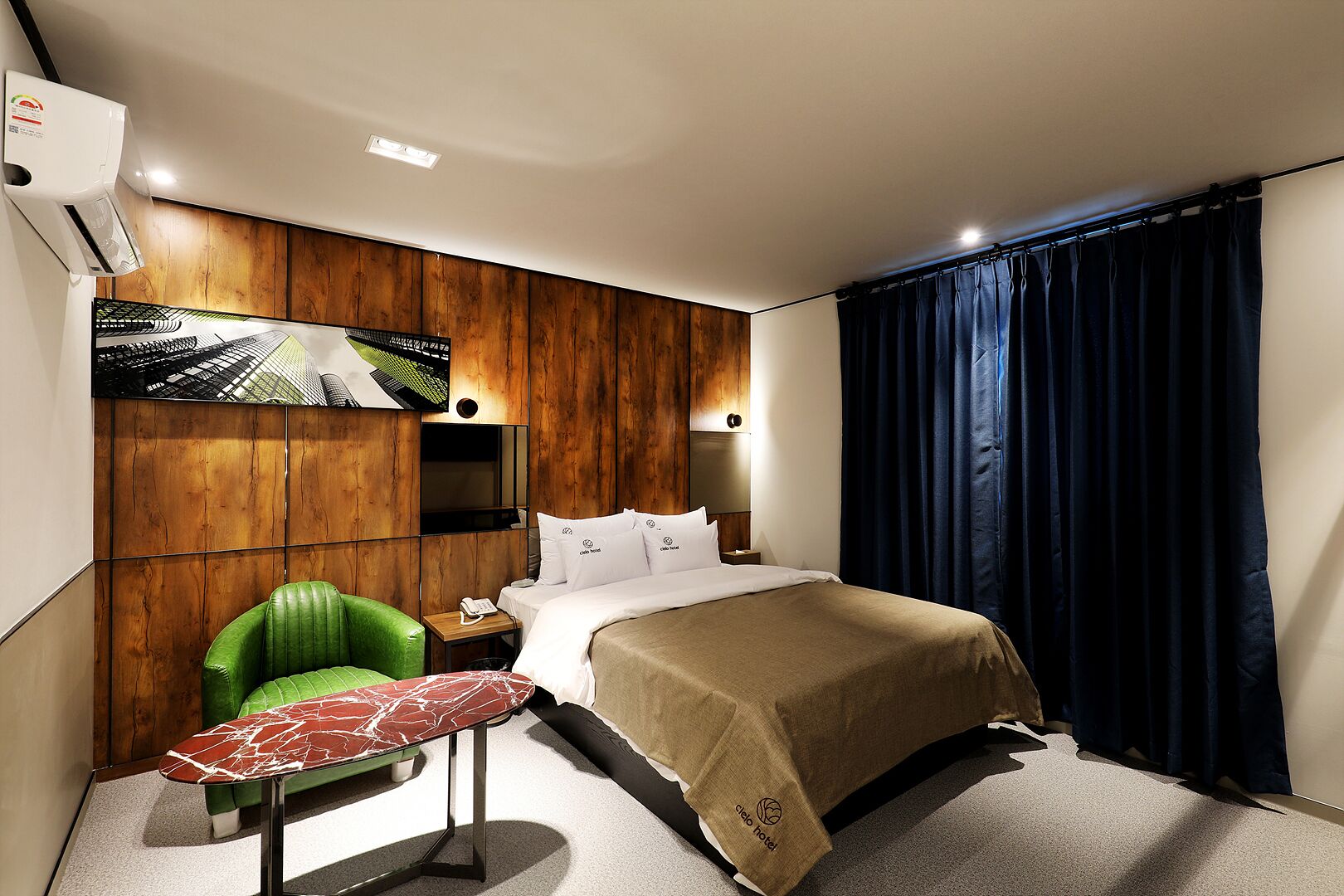 A hotel room with a bed, a green chair, a small table, a TV mounted on the wall, and a window with dark curtains.