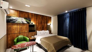 A hotel room with a bed, a green chair, a small table, a TV mounted on the wall, and a window with dark curtains.