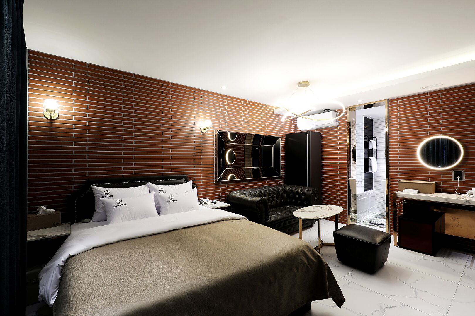 Habitación de hotel moderna con pared de ladrillo, una cama grande con ropa blanca, un sofá de cuero, una mesita redonda pequeña, un tocador con espejo y un armario empotrado.