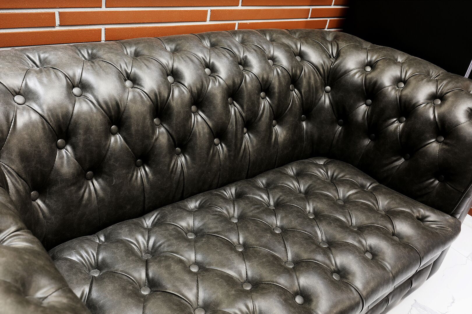 A dark brown leather sofa with a tufted design against a brick wall.