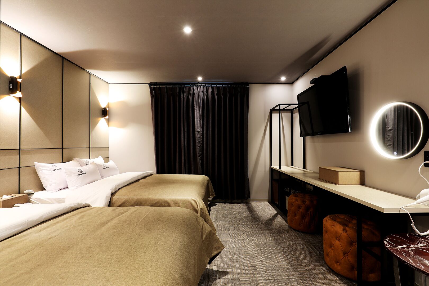 A modern hotel room with two beds, a desk with a flat-screen TV, a round mirror, and a small seating area.
