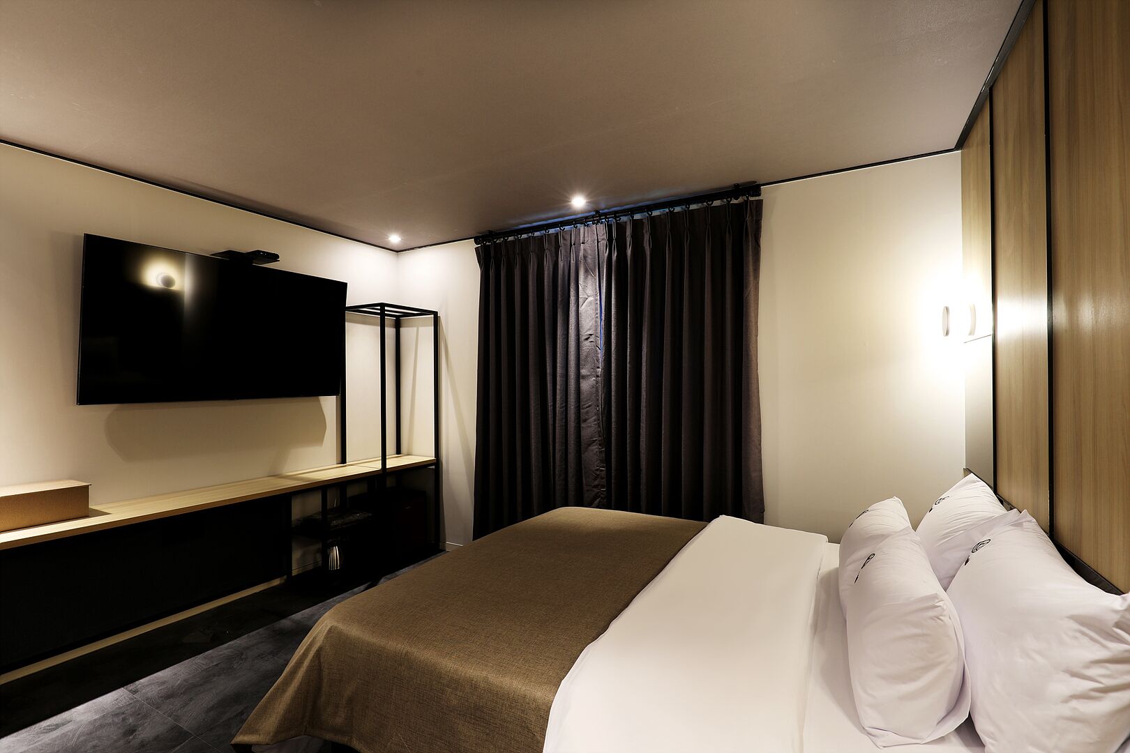 A modern hotel room with a flat-screen TV mounted on the wall, a wooden bedside table, a bed with white linens, and dark curtains.