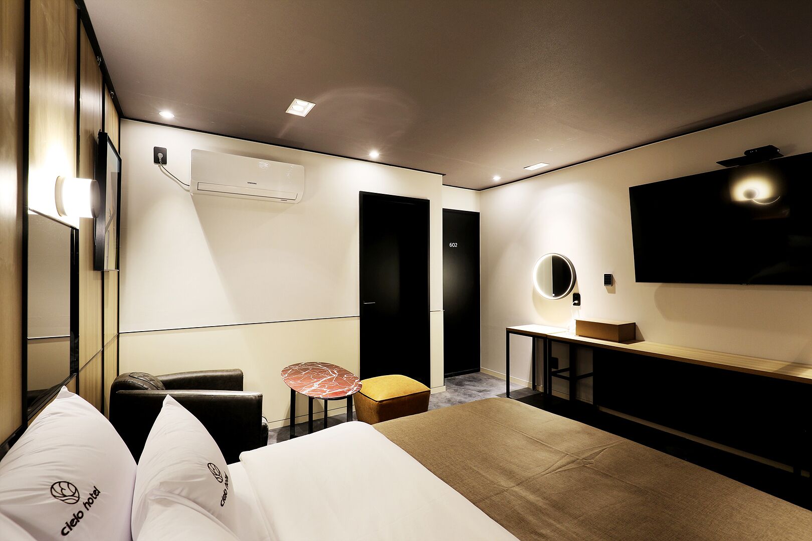 Quarto de hotel moderno com cama, escrivaninha, cadeira, mesa pequena, espelho e televisão.