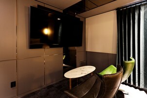 Una habitación moderna con un televisor de pantalla plana montado en la pared, una mesa redonda con tapa de mármol y dos sillones de cuero con cojines verdes.