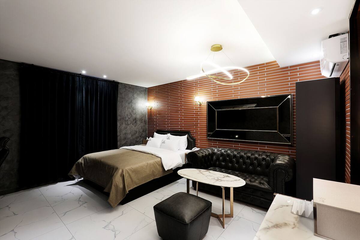 A modern hotel room with a bed, a black leather sofa, a small round table, and a TV mounted on the wall.