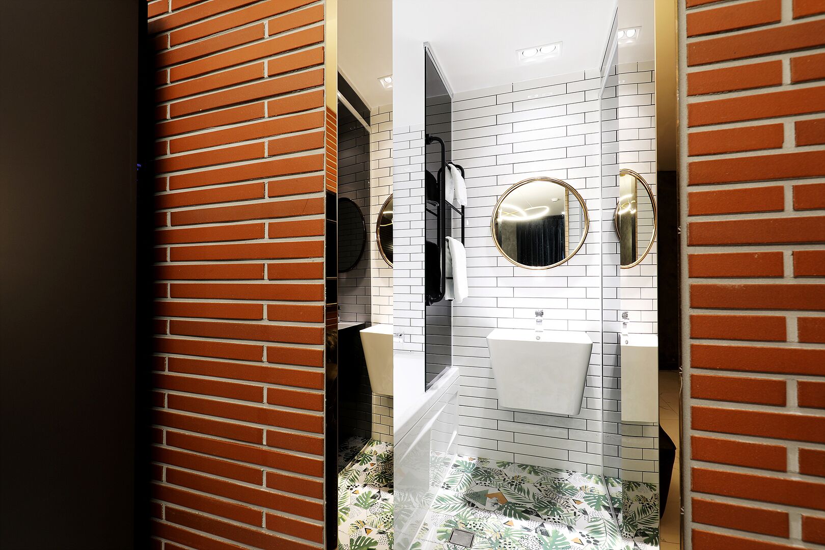 Un baño moderno con pared de ladrillo, azulejos blancos tipo metro, un espejo redondo y un lavamanos blanco.