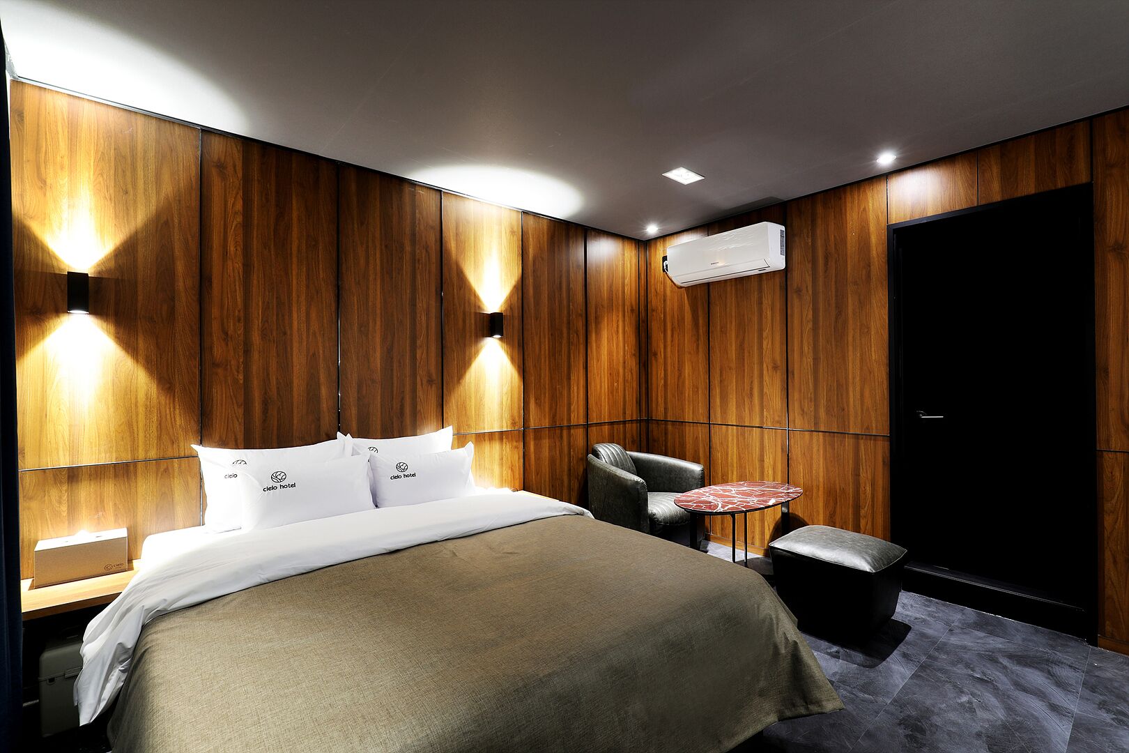 A hotel room with a bed, a chair, a small table, and an air conditioning unit.