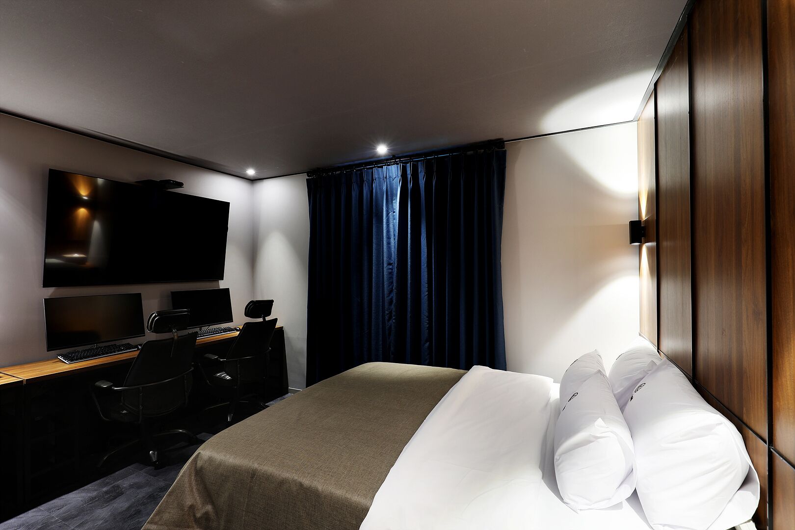 A modern hotel room with a large bed, a desk with a computer, a flat-screen TV, and a dark curtain.