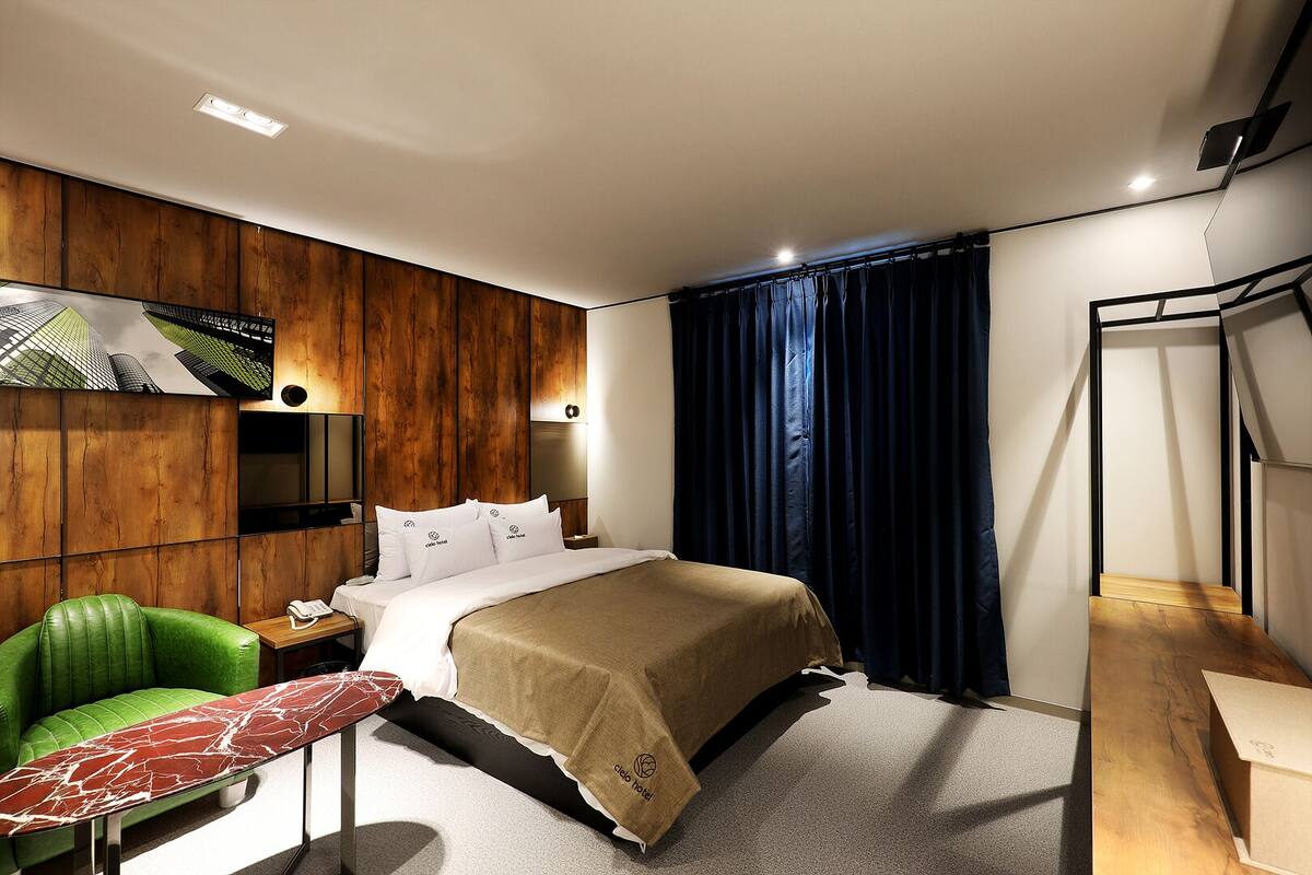 A modern hotel room with a bed, a green chair, a small table, and a TV mounted on the wall.