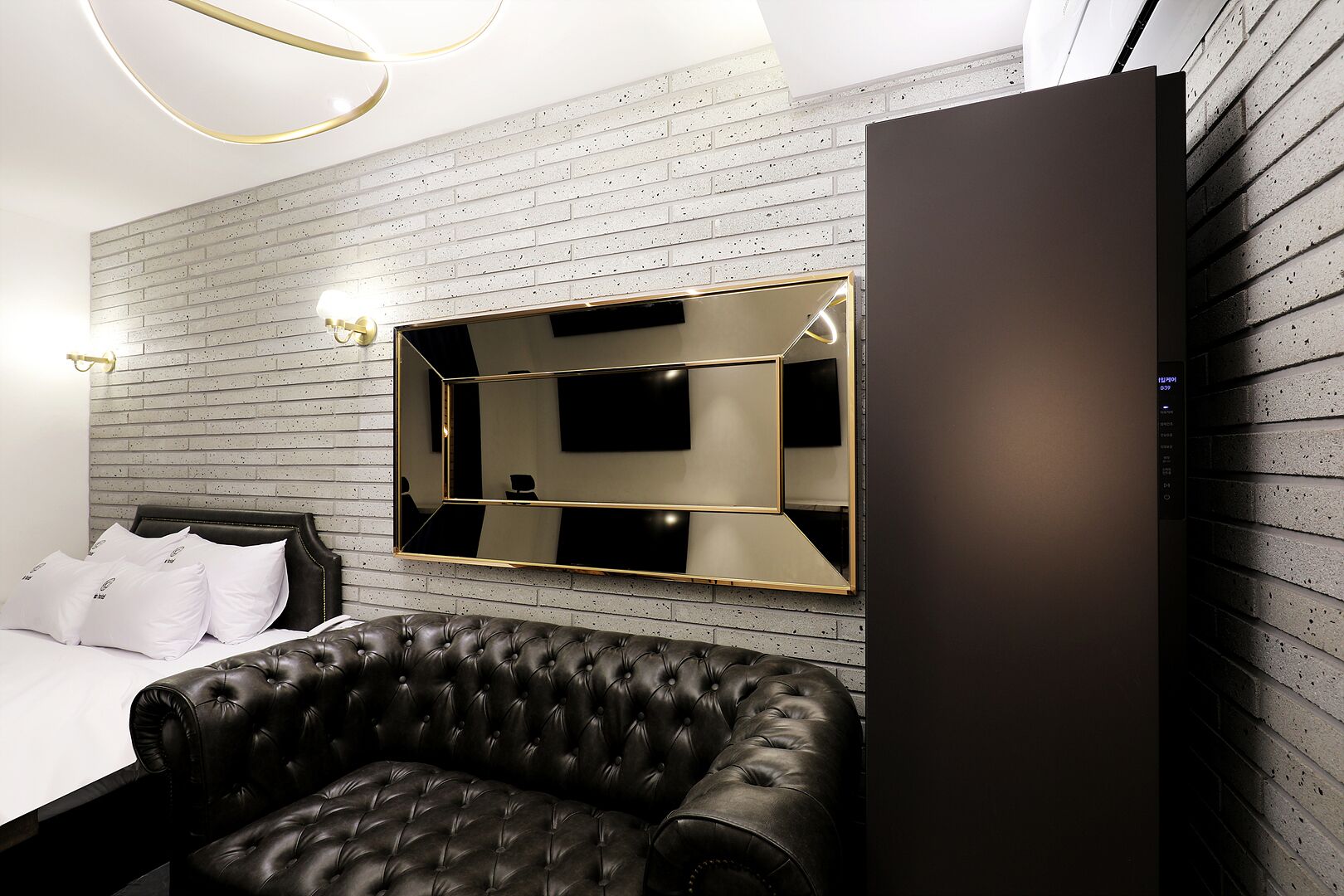 A modern hotel room with a bed, a black leather sofa, a large mirror, and a wall-mounted TV.