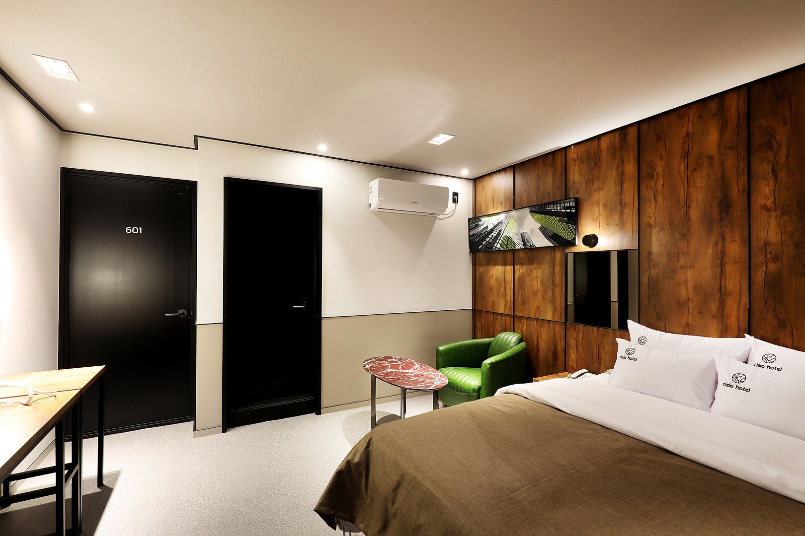Una habitación de hotel moderna con una cama, una silla verde, una mesita y un televisor en la pared.