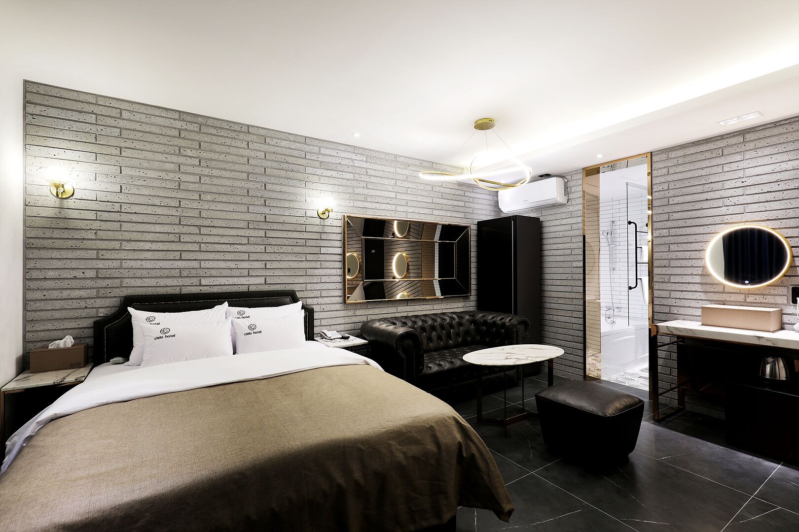 Une chambre d’hôtel moderne avec un mur au motif de briques, un grand lit avec du linge de lit blanc, un canapé en cuir noir, un miroir rond et une petite table ronde.
