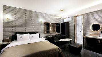 Une chambre d’hôtel moderne avec un mur au motif de briques, un grand lit avec du linge de lit blanc, un canapé en cuir noir, un miroir rond et une petite table ronde.