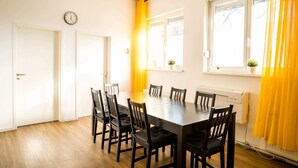 A dining area with a rectangular table surrounded by chairs, a clock on the wall, and windows with curtains. - Spacious 3 Bedroom Apartment Online Check-In (Berlin)