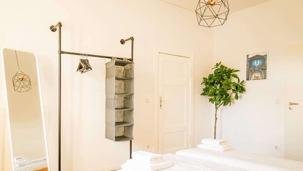 A hotel room with a bed, a wardrobe, a mirror, a hanging rack, a plant, and a door. - Spacious 3 Bedroom Apartment Online Check-In (Berlin)