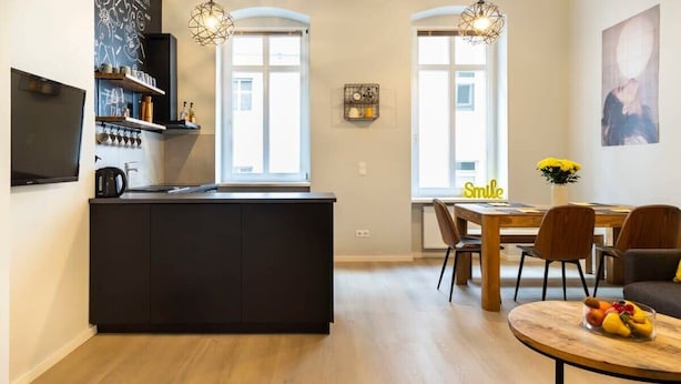 Een moderne keuken met een donker keukenblok, houten vloeren en hangende lampen. Er is een eethoek met een tafel en stoelen, en een tv aan de muur.