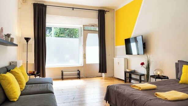 Moderni hotellihuone, jossa on harmaa sohva, harmaalla päiväpeitteellä varustettu sänky, taulutelevisio ja suuri ikkuna, jossa on verhot.