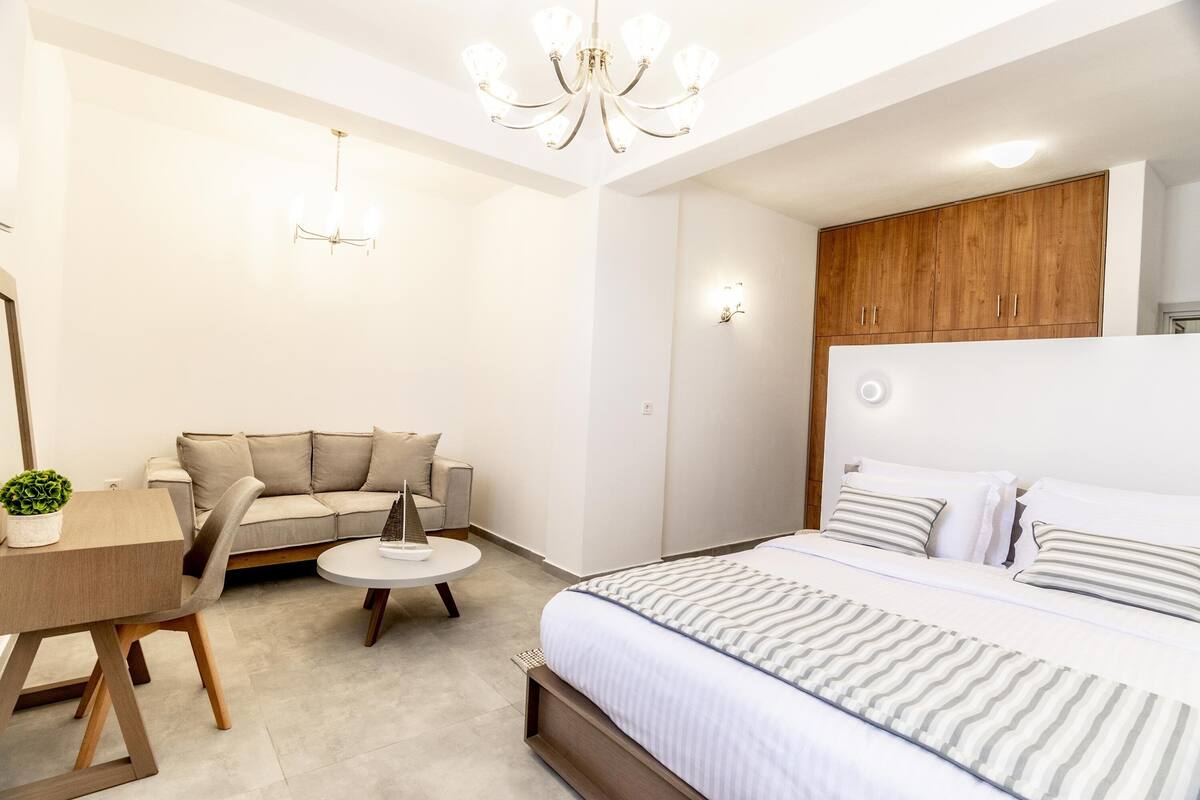 A modern bedroom with a large bed, a wooden headboard, a round coffee table, a sofa, and a desk with a plant.