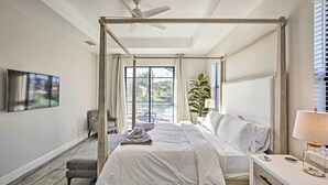Un dormitorio moderno con cama con dosel, televisor de pantalla plana, escritorio con lámpara y vistas a una piscina y zonas verdes.