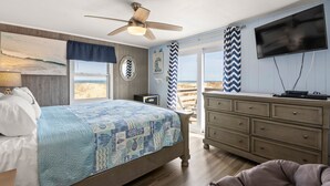 A bedroom with a bed, a dresser, a television, and a view of the beach. - Windjammer: Oceanfront, Shared Pool, Beach Access (Nags Head)