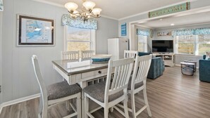 A dining area with a table, chairs, and a chandelier. There is a refrigerator and a television in the background. - Windjammer: Oceanfront, Shared Pool, Beach Access (Nags Head)
