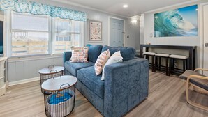 A living room with a blue sofa, two round tables, a large window with blinds, a door, and a wall-mounted bar with stools. - Windjammer: Oceanfront, Shared Pool, Beach Access (Nags Head)