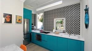 A modern kitchen with teal cabinetry, a stainless steel refrigerator, and a window with a geometric patterned wall.