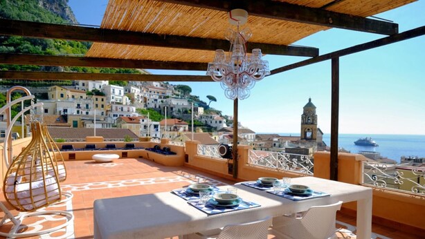 A terrace with a table set for two, overlooking a coastal town with a church and a ship in the distance.