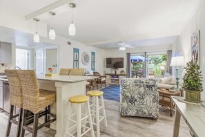 Amplia sala de estar con cocina, comedor y un televisor grande. Hay sillas de mimbre, un taburete de bar y un sofá estampado.