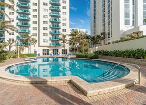 Piscina com borda de tijolos, cercada por palmeiras e um prédio alto ao fundo.