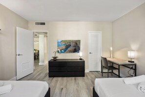 A hotel room with two beds, a desk, a dresser, and a painting of a coastal landscape.