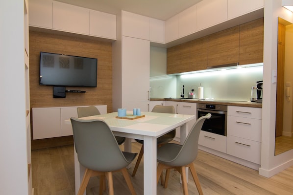 A modern kitchen with a flat-screen TV, wooden cabinets, and a dining area with chairs and a table.
