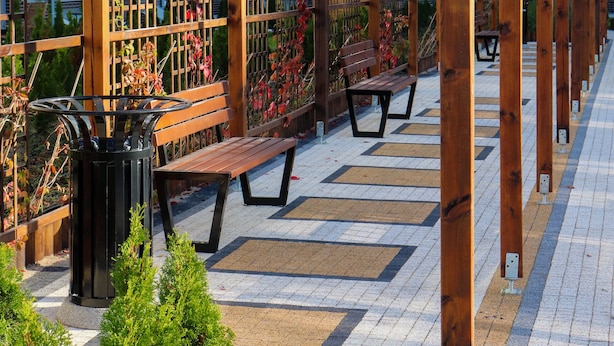 Une pergola en bois, dotée de bancs et d’une poubelle, se trouve dans un espace extérieur dont le sol est recouvert de carreaux et qui est agrémenté de verdure.