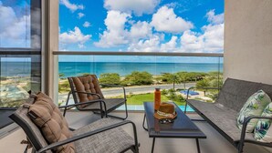 A balcony with a view of the ocean and a resort area. There are two chairs and a table with a hat and a bottle on it. - Discover Ocean View Condo Living (Oranjestad)